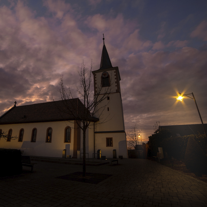 Das Bild zeigtdie alte Kirche von Waldbüttelbrunn im Abendlicht, Straßenlaternen leucht